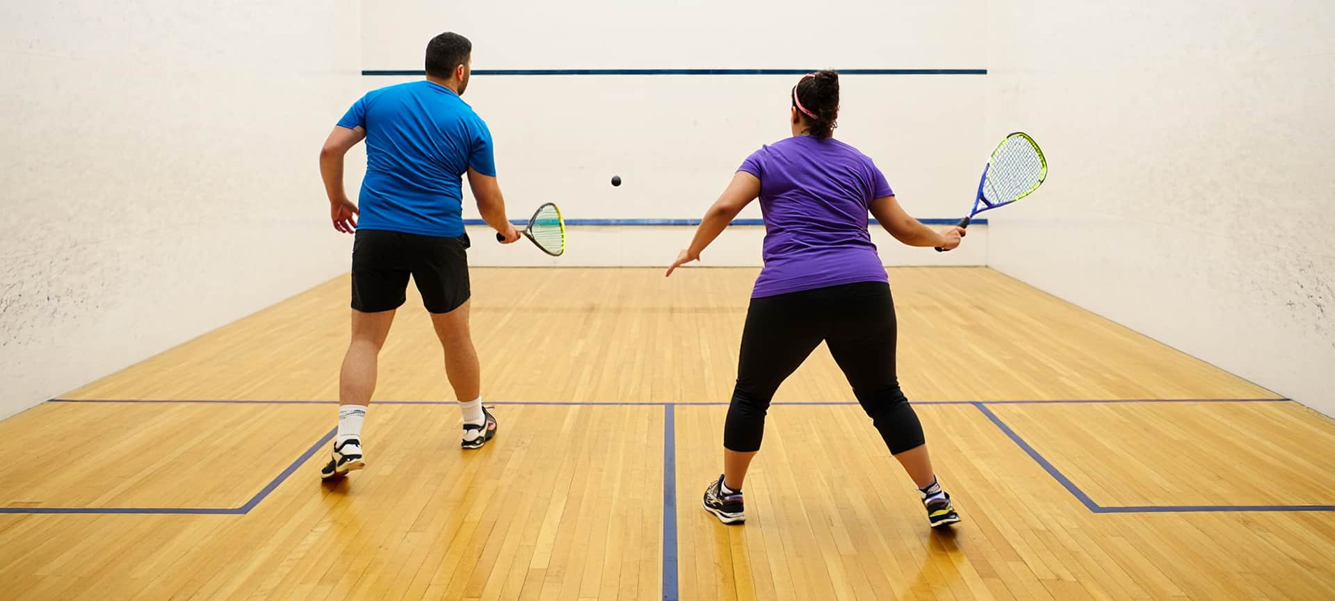 Man And Woman Playing Racquetball On An Indoor Court, Both Holding Rackets And Facing The Front Wall.